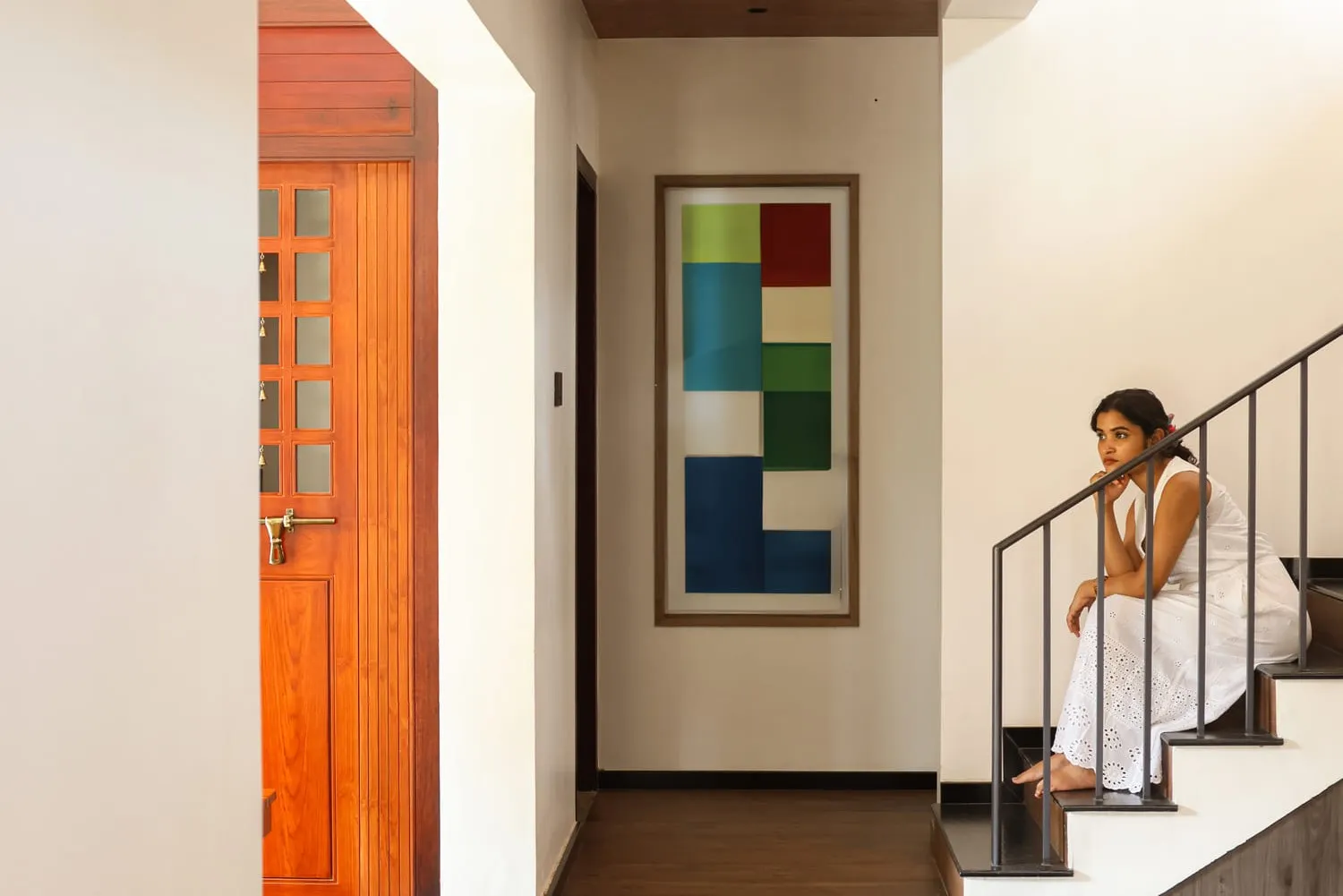 Interior hallway with a wooden door, colorful abstract painting, and a person sitting on stairs.