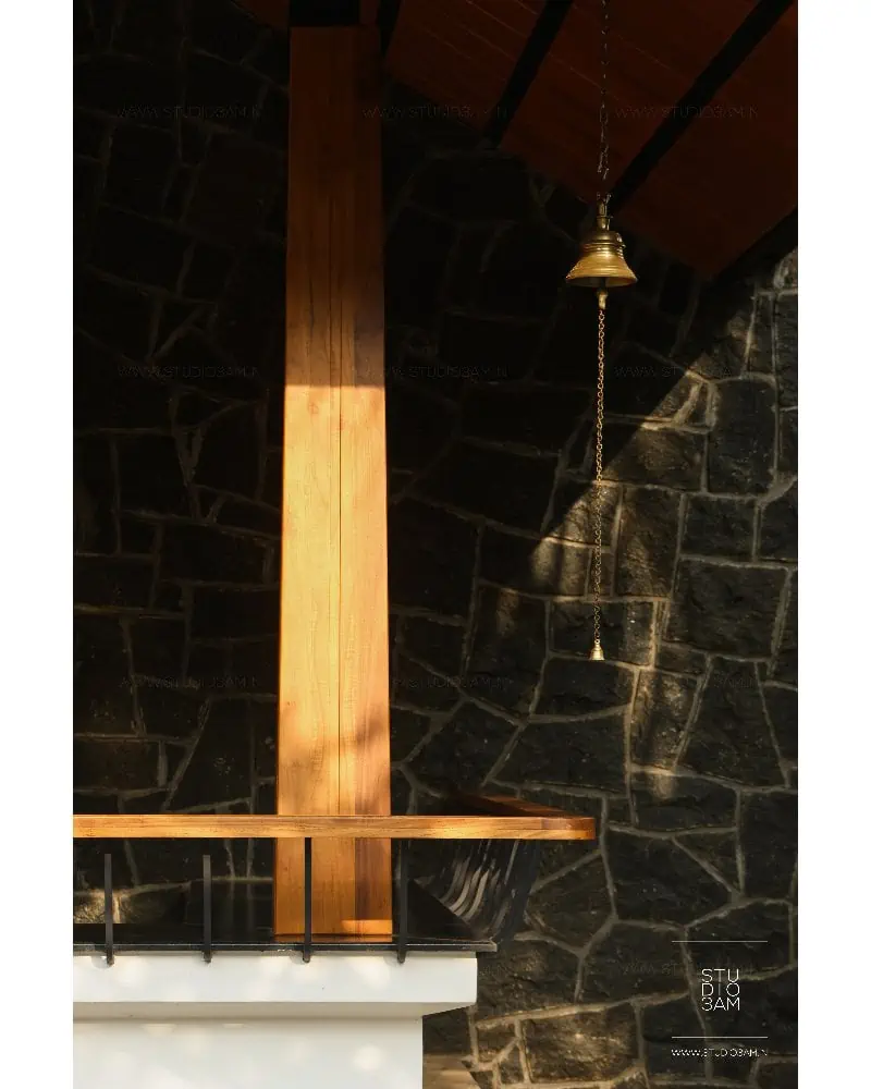 A cozy interior features a wooden beam extending up a stone wall with a hanging brass bell. The wall is constructed of dark, irregularly shaped stones, and a wooden countertop is visible in the foreground. Warm, ambient lighting enhances the rustic feel.