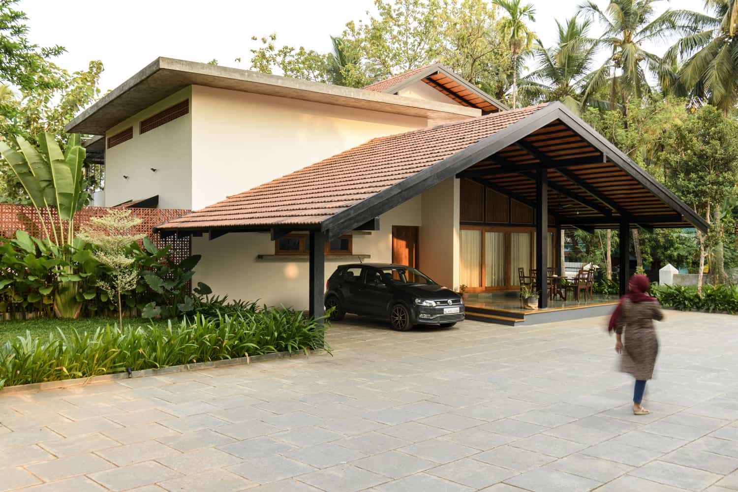 A modern house with a red-tiled roof and an angled carport. A black car is parked under the carport. A person in a maroon headscarf and gray coat walks on the stone-paved driveway. The house is surrounded by lush greenery and tall trees.