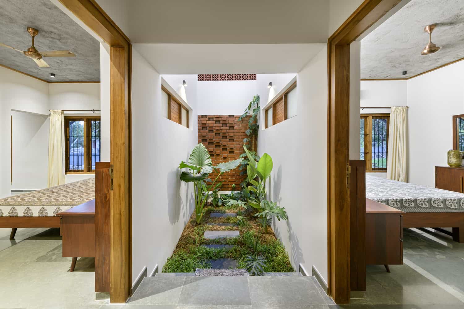 A modern indoor space featuring two bedrooms on either side of a hallway with large plants and greenery. The hallway features a small indoor garden with various plants and stepping stones. Wooden furniture, ceiling fans, and large windows are seen in both bedrooms.