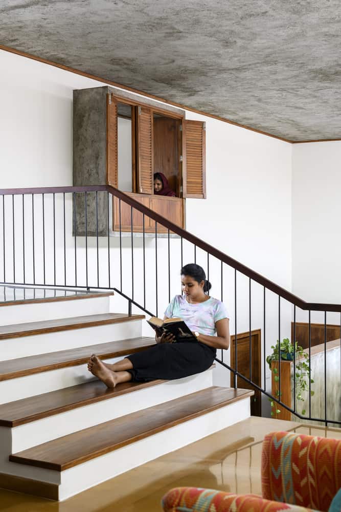 A woman sits on indoor stairs, reading a book. She leans against the railing, and in the background, another woman is partially visible, looking through a small window with wooden shutters inside the house. There are plants and wooden furniture around.