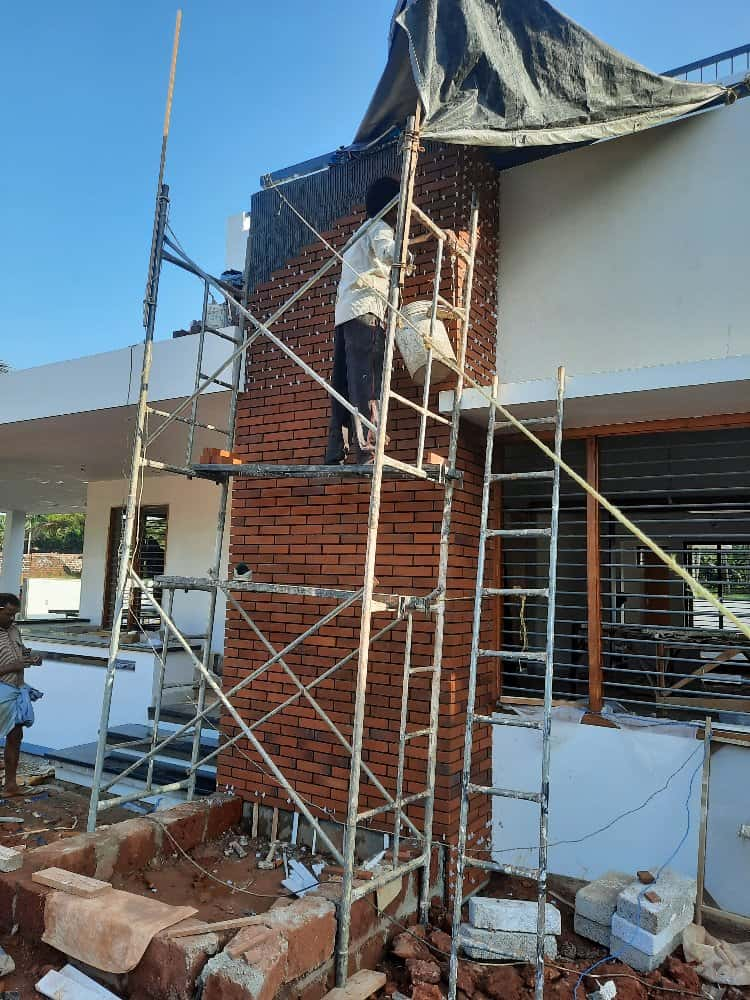 A construction worker stands on scaffolding, working on a brick façade of a building. The area is partially covered with a tarp. 