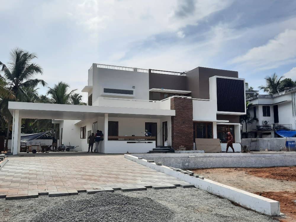 A modern two-story house with a flat roof, featuring white and brown exterior walls. Construction materials and workers are evident on the premises.