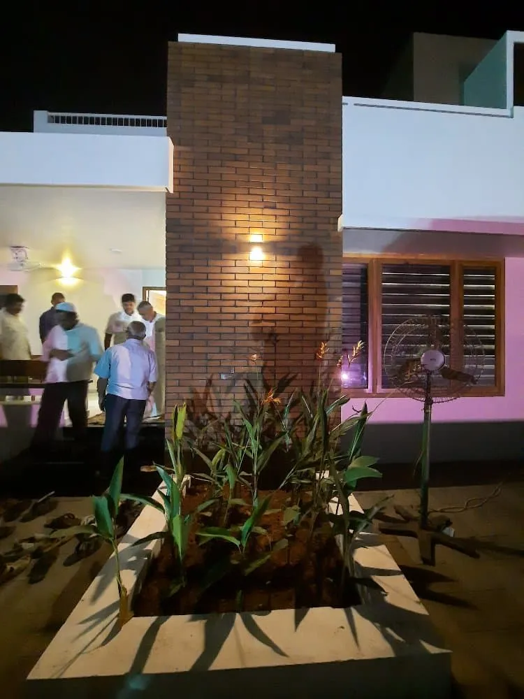 A group of people stand outside a modern white building with a brick column feature illuminated by a light. In the foreground, there is a rectangular planter with leafy green plants and a fan nearby. 