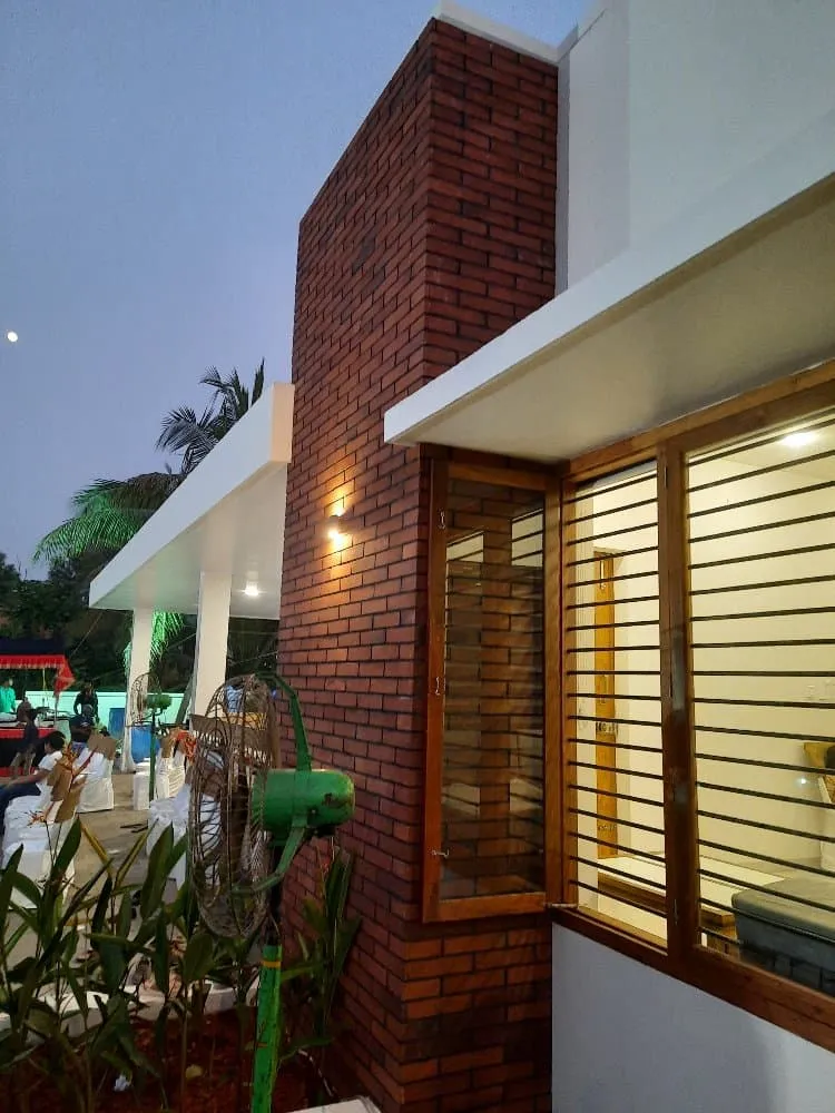 A modern brick and white building facade featuring large windows with horizontal bars, illuminated by an exterior light at dusk. Plants and a green fan are in the foreground, and people are visible in the background under a covered patio.